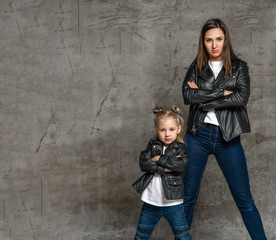 Positive young woman and little girl in similar jeans and black leather jackets standing in same...