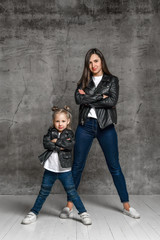 Young mother and little daughter are posing against gray studio background. Wearing in black leather jackets, white t-shirts, blue jeans. Close up