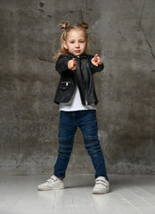 Obraz na płótnie Canvas Little cute blond girl in stylish rock style black leather jacket, jeans and sneakers standing and pointing at camera with fingers