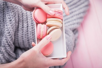Female hands holding rectangular white plate with delicious pink and beige macaroons on pink and gray background. Top view, copy space