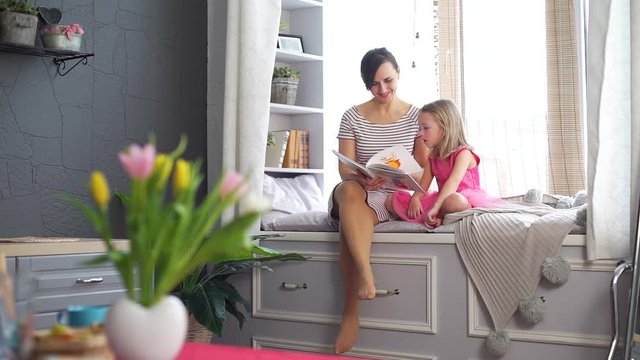 Young Woman Reading Book with Little Daughter at Home. They Sitting on the Big Cozy Windowsill. Slow Motion. People, Lifestyle and Happy Family Concept