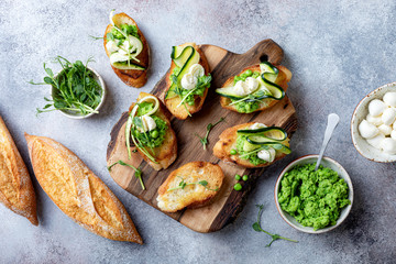 Appetizer crostini with mashed green pea, mozzarella, pea sprouts and zucchini ribbons on wooden board. Delicious healthy snack, spring appetizers