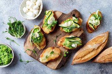 Appetizer crostini with mashed green pea, mozzarella, pea sprouts and zucchini ribbons on wooden board. Delicious healthy snack, spring appetizers