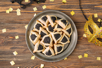 Plate with tasty hamantaschen and party decor for Purim holiday on wooden background