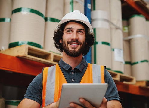 Successful warehouse supervisor smiling and working while holding digital tablet to check inventory and stock wearing white helmet and safety jacket