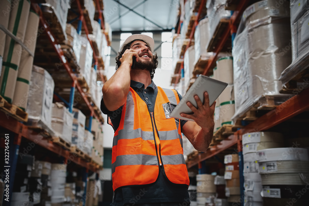 Wall mural low angle view of warehouse manager talking over phone while smiling and holding digital tablet stan