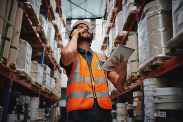 Low angle view of warehouse manager talking over phone while smiling and holding digital tablet standing in aisle with goods