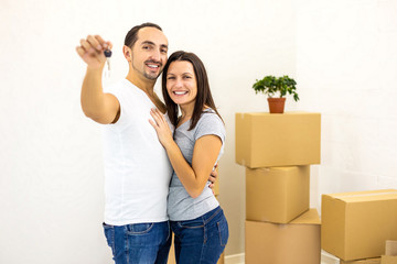 Lovers standing together on white background with piles of boxes, man showing keys to their new house.