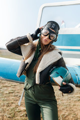 a girl in a headdress and a pilot s glasses holds a helmet in her hands, is dressed in a green jacket, a green overalls, against the background of the wing and door of the plane