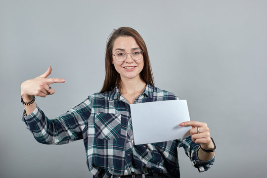 Young brunette girl blue green in checked shirt on grey background a happy woman with glasses holds an empty white sheet of paper in hand, points at it with index finger
