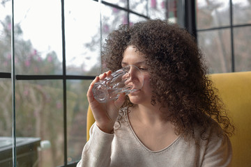 Young woman with curly hair is sitting in a cafe and drinking water