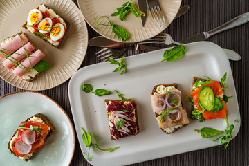 Selection of Danish smorrebrod open sandwiches on platter and on plates, cutlery on gray textile background, top view