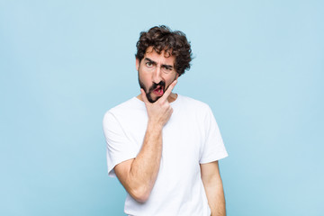 young crazy bearded man with mouth and eyes wide open and hand on chin, feeling unpleasantly...