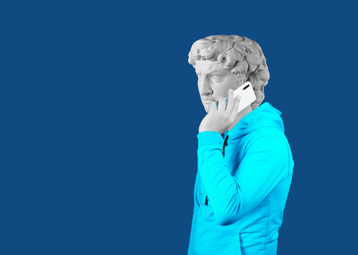 Modern art collage. Concept portrait of a man holding mobile smartphone using app texting sms message. Gypsum head of of David. Man in suit. On a blue background.