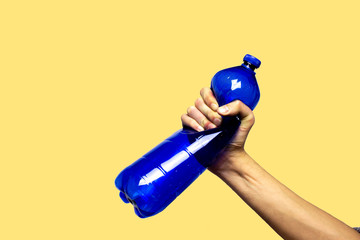 Concept of stop plastic pollution, global warming, recycling plastic, plastic free. Hand tightly squeezes an empty blue plastic bottle in a sign of protest. Yellow background with an isolated subject