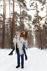 Fototapeta premium Happy lovers a guy and a girl who love each other hug, kiss, laugh, rage and walk in warm jackets in winter against the background of a snowy forest, a friendly family fun walk