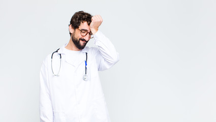 young doctor man raising palm to forehead thinking oops, after making a stupid mistake or remembering, feeling dumb against copy space wall