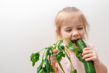 a little girl in a light T-shirt holds a bunch of parsley in one hand, in the second a big cucumber, bites off a cucumber and laughs