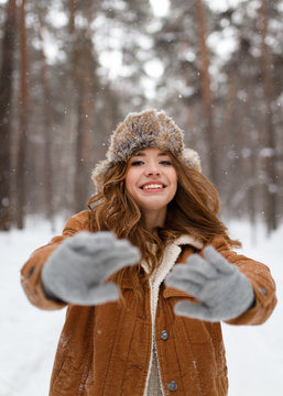 A beautiful cute cheerful young girl with curly hair and a snow-white smile in a brown jacket and fur hat walks and plays with snow in the winter forest against the background of trees, enjoy