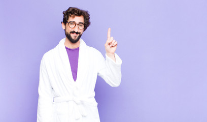 young bearded man wearing bathrobe smiling cheerfully and happily, pointing upwards with one hand...
