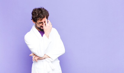 young bearded man wearing bathrobe looking stressed, ashamed or upset, with a headache, covering...