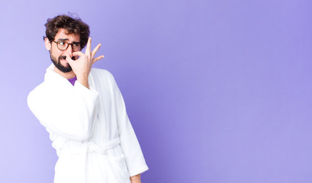 young bearded man wearing bathrobe feeling disgusted, holding nose to avoid smelling a foul and unpleasant stench