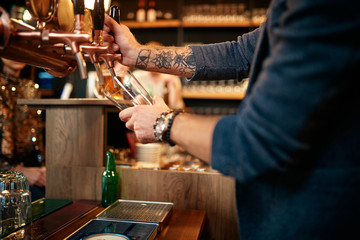 Tattooed caucasian barman pouring beer while standing in pub. Selective focus on hand.