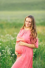 Fototapeta na wymiar pregnant woman in pink dress touches belly and looks into camera