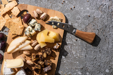 Top view of pieces of cheese, olives, pistachios, slices of pear, crackers and knife on cutting board on grey background
