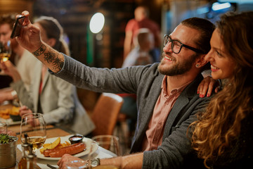 Cute caucasian smiling couple in love sitting in restaurant and taking selfie. Around them are their friends chatting and having dinner.