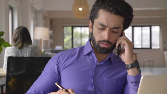 a young Indian male entrepreneur busy over a conversation on a cell phone sitting right in front of a desk noting down something while operating laptop at his workplace