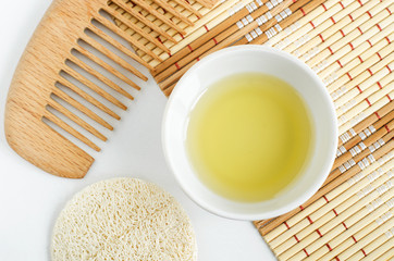 Small white bowl with olive (or coconut) oil, loofah sponge and wooden hair comb. Homemade skin and hair treatment concept. Top view, copy space