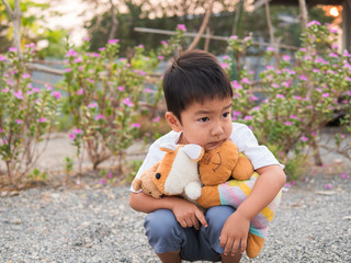 Asian little child boy holding, hugging favorite teddy bear with serious face. Kid in white t-shirt thinking, in rural natural background.