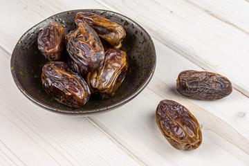 Lot of whole dry brown date fruit in dark ceramic bowl on white wood