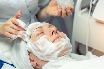 Close-up beautician doctor hands making anti-age procedures, applying foam cleansing mask for mid-aged female client at beauty clinic. Cosmetologist doing skincare treatment .Health care therapy