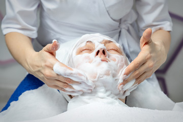 Close-up beautician doctor hands making anti-age procedures, applying foam cleansing mask for mid-aged female client at beauty clinic. Cosmetologist doing skincare treatment .Health care therapy