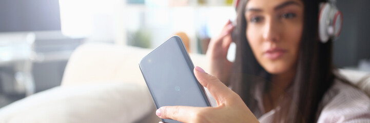 Young beautiful woman headphones holds smartphone in hand using at home costs about sofa. makes...