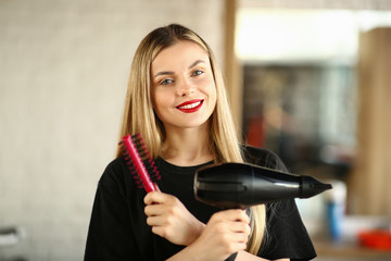 Fototapeta premium Smiling Hairstylist with Hairdryer and Round Comb. Woman Hairdresser Holding Black Blow Dryer and Hairbrush for Styling Haircut. Beautiful Stylist with Equipment for Haircut Looking at Camera Shot