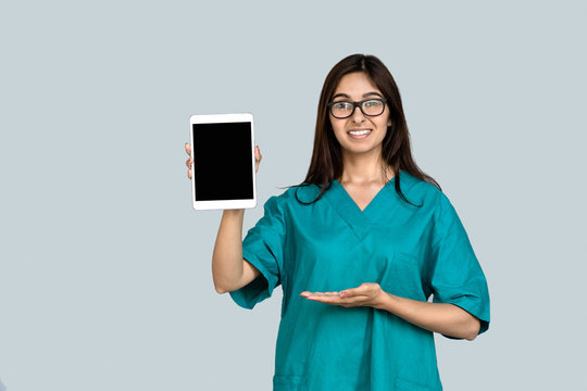 Happy young indian woman professional nurse female doctor wear green uniform show digital tablet mock up blank empty screen advertise medical care app tech concept isolated on gray studio background.