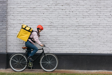 Side view of smiling courier with thermo backpack riding bicycle near building