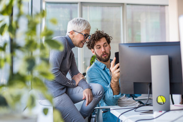 Two co-workers talking on a work break at the office. The man is showing something to the woman on his phone while she sits leaning against the window sill.