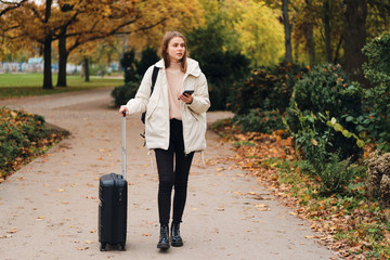 Fototapeta premium Attractive casual girl in down jacket with suitcase and cellphone walking through park