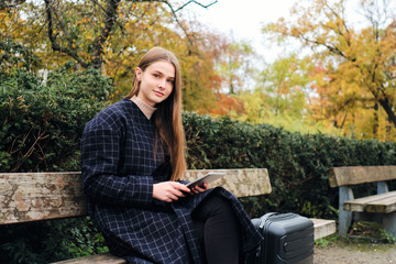 Attractive casual girl using tablet sitting with suitcase on bench in park