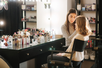 Beautiful female makeup artist doing makeup for a young blond hair girl in a beauty salon sitting...