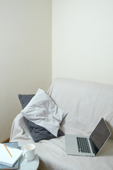  An open computer is on the sofa next to the pillows. On the table is a cup, notebook and pencil. Freelancer workplace concept
