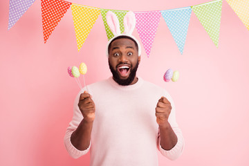 Photo of funny dark skin guy visit family easter party hold festive colorful painted eggs on sticks wear sweater pants bunny ears dotted flags hang pink color background