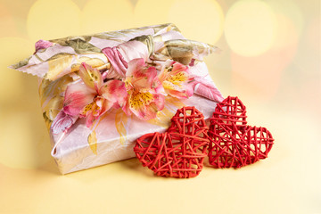 Trendy wrapped in Furoshiki technique gift box with flowers Alstroemeria and two red wicker hearts on yellow with bokeh.