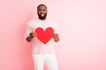 Photo of attractive dark skin guy hold red paper heart affectionate boyfriend inviting girlfriend romantic date wear knitted sweater white pants isolated pastel pink color background