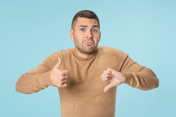Young man confused with thumbs up and down trying to take a decision over blue background