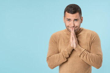 Young handsome man asks for something keeps palm together over isolated blue background